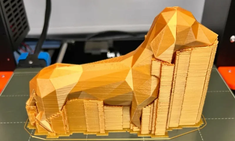 Un modelo de perro impreso en 3D en color dorado con diseño geométrico y estructuras de soporte visibles en una cama de impresora.