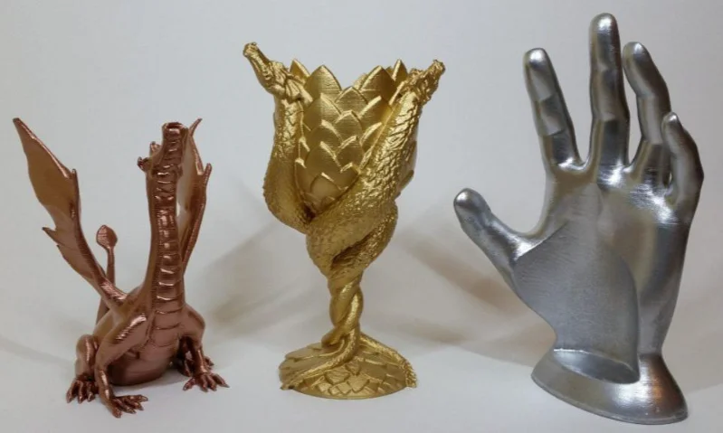 Tres objetos impresos en 3D: un dragón de bronce, una copa de oro y una mano de plata.