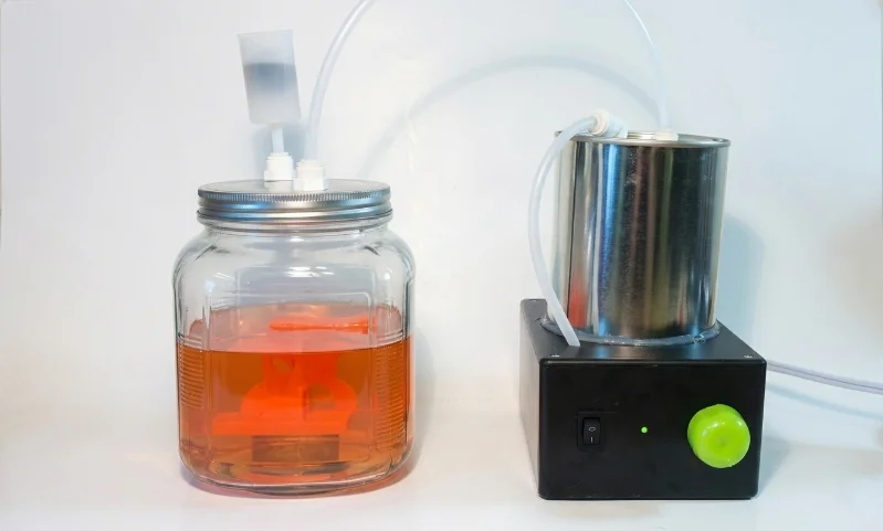 Configuración de anodizado de bricolaje con un recipiente de vidrio con líquido naranja y un dispositivo metálico conectado mediante tubos para el proceso de anodizado.