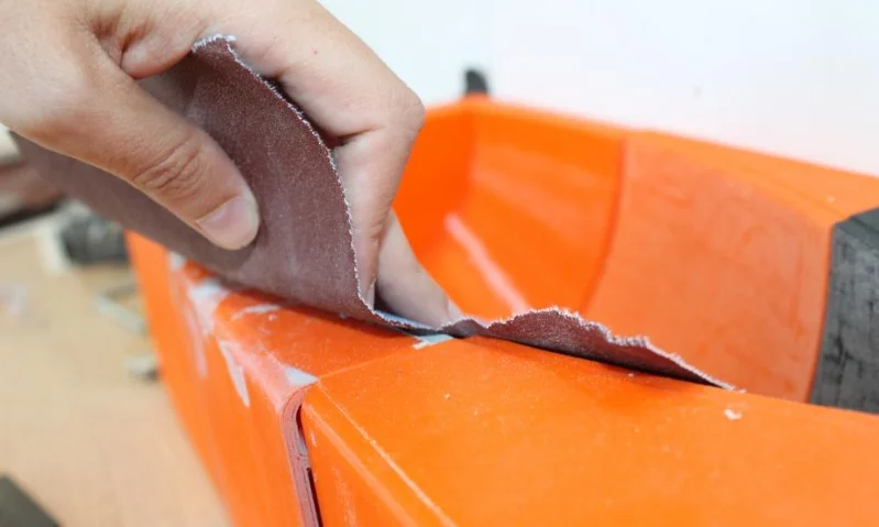 Mano sosteniendo papel de lija para alisar el borde de una superficie de plástico naranja durante el acabado.