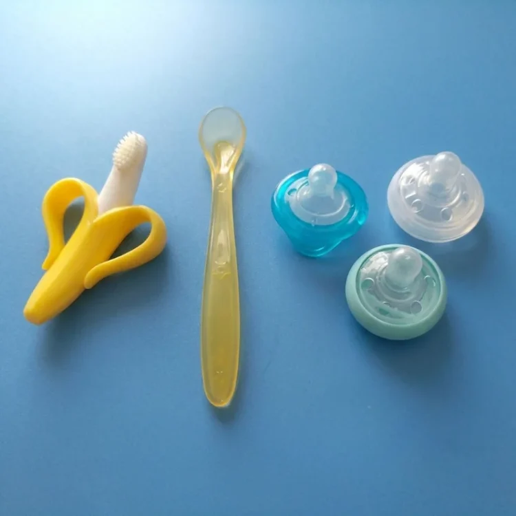 Yellow banana-shaped toothbrush, silicone baby spoon, and three pacifiers on a blue surface.